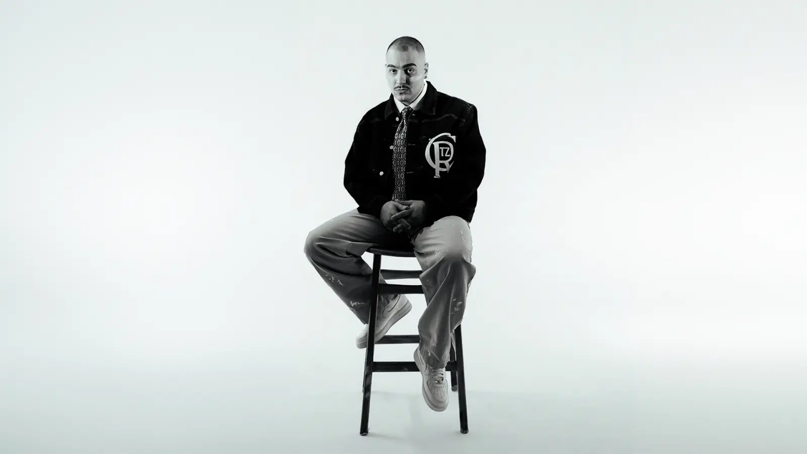 JEFE tour photo: the Turkish rapper in a black varsity jacket and grey trousers, sitting on a wooden stool against a plain white background