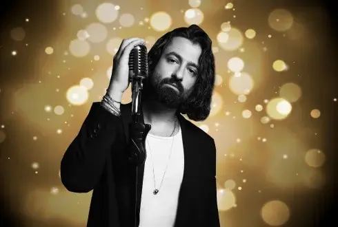 Kaya Hotels artist photo: Koray Avcı with a beard in a black jacket against a gold bokeh background
