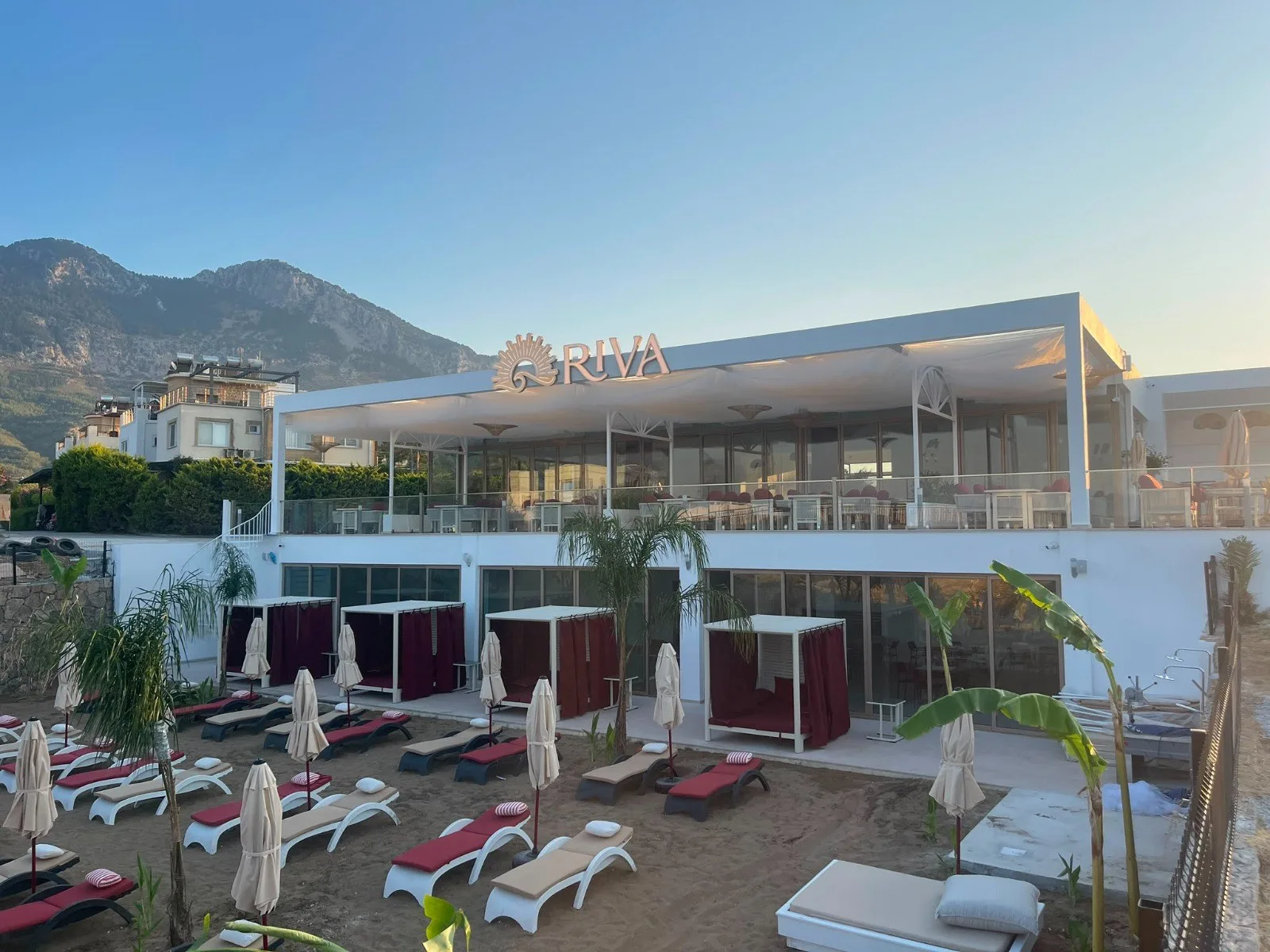 RIVA Beach Club building in Lapta with red-curtained cabanas and sunbeds on the sand, mountains behind
