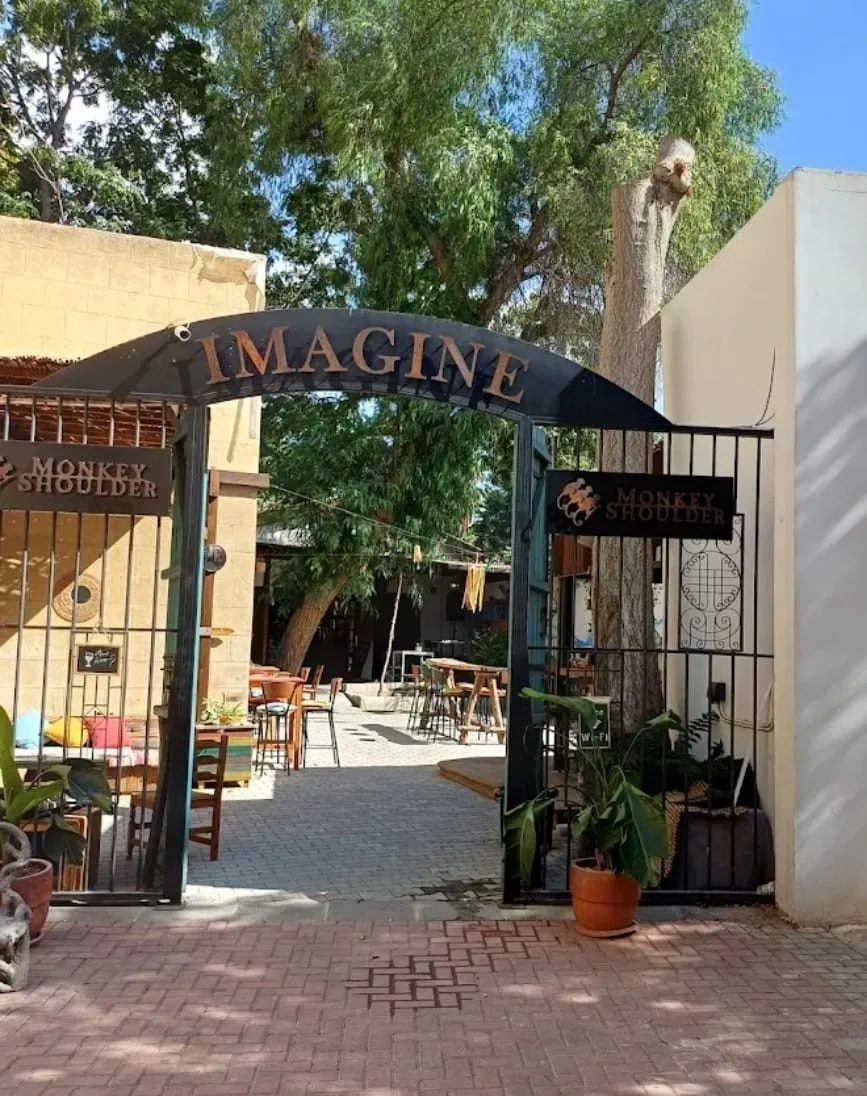 Arched IMAGINE sign over the open gate of Imagine Cafe Bar, with a paved courtyard and plants behind