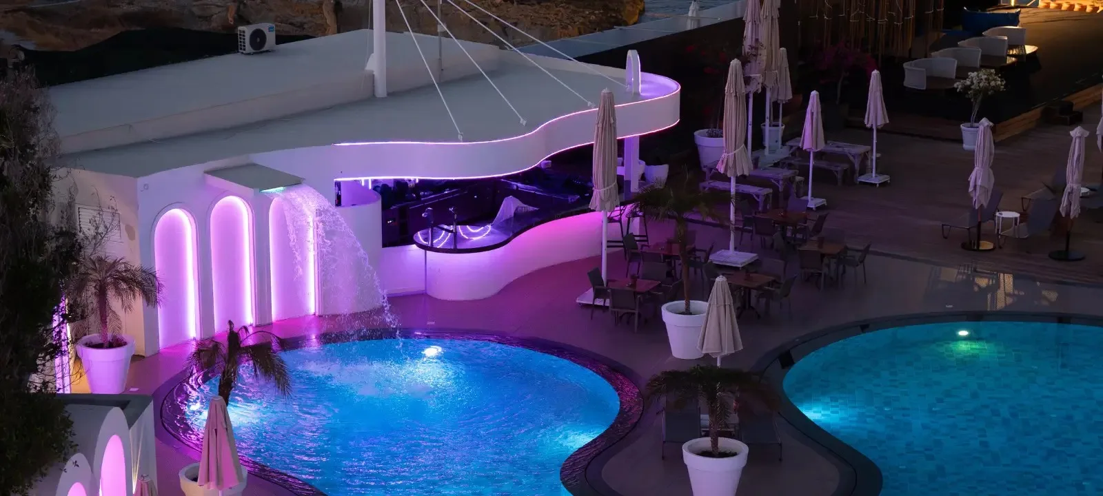 Byblos at Rocks Hotel at night: a white curved bar lit in purple beside two illuminated pools