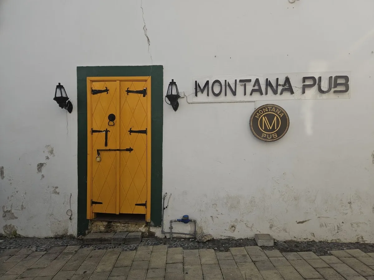 Girne Ağa Cafer Paşa Sokak'taki Montana Pub girişi: sarı ahşap kapı, duvar fenerleri ve beyaz duvarda Montana Pub tabelası