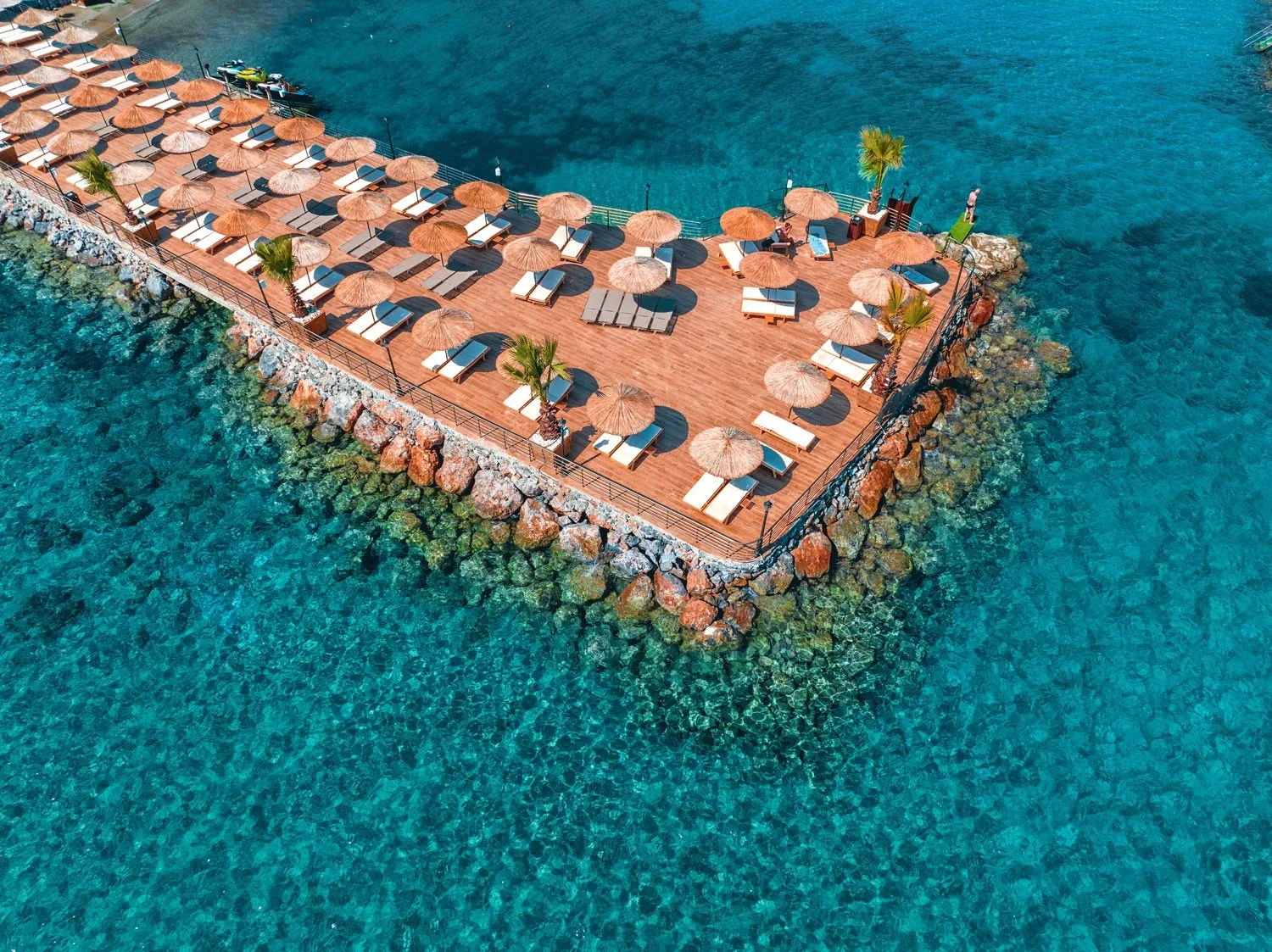 Aerial view of Suna's Beach Club's wooden sun deck with straw umbrellas and sunbeds over turquoise water