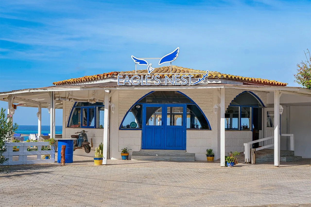 Exterior of Eagle's Nest in Tatlısu: white seaside building with blue doors and EAGLE'S NEST roof sign, sea behind