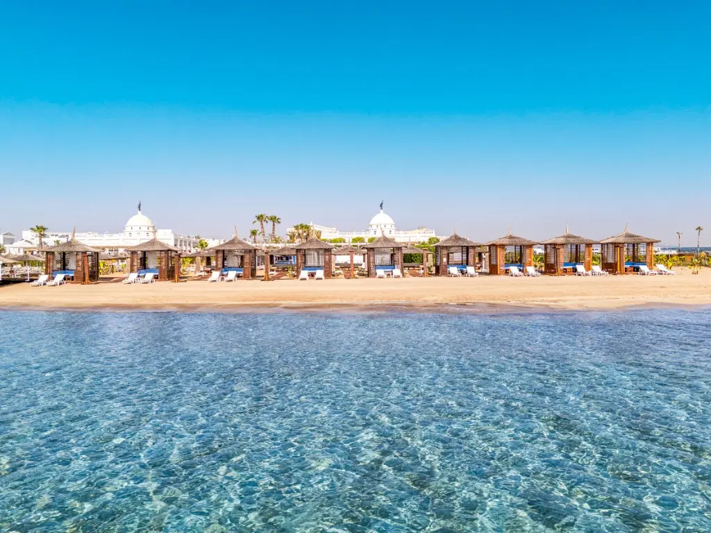 Kaya Artemis Resort beach with wooden cabanas and clear sea, white domed hotel buildings behind, Bafra, İskele