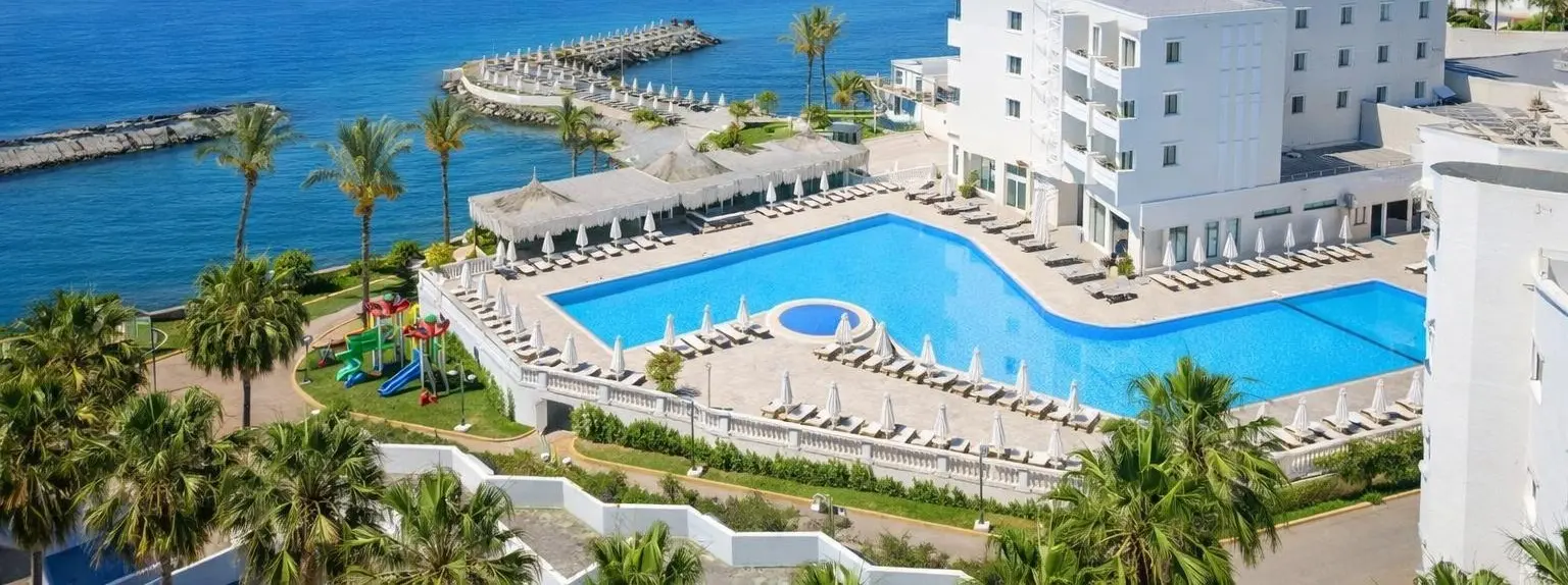 Aerial view of Vuni Palace Hotel in Kyrenia: white hotel blocks beside a large pool with sun loungers, palm gardens and the sea and pier behind